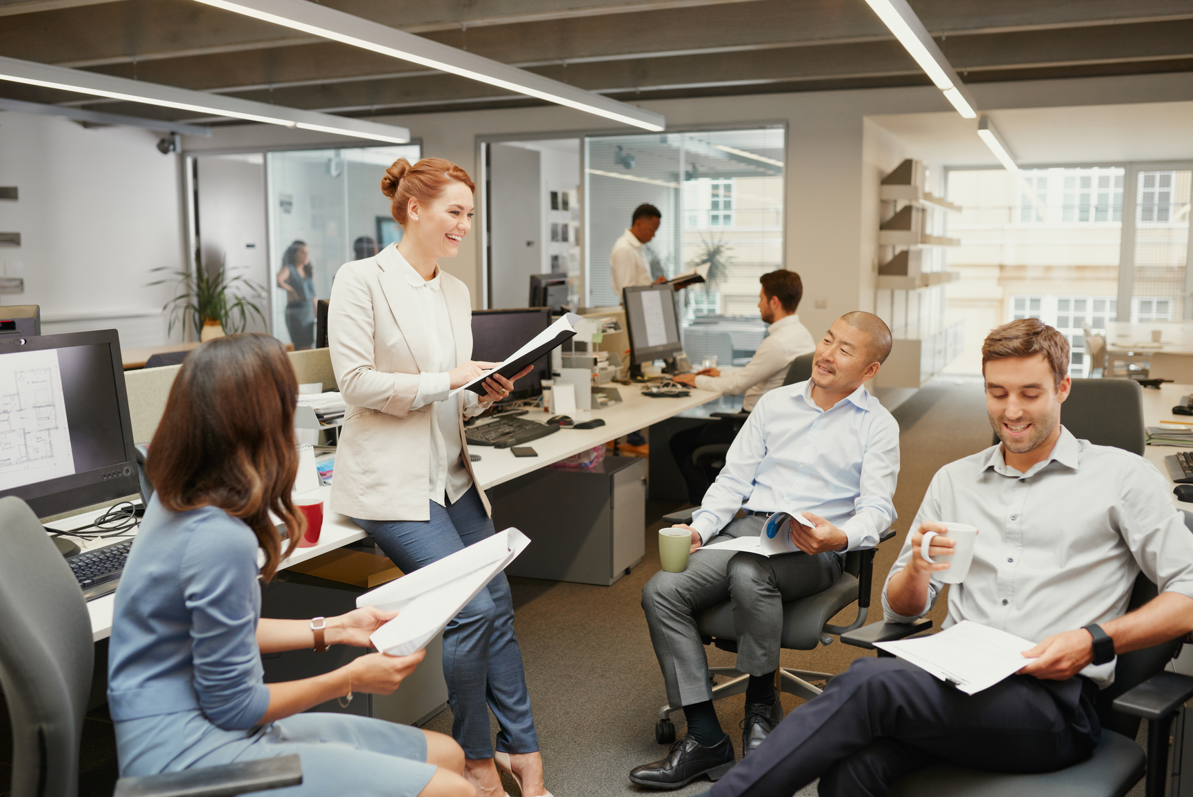 Attractive redhead woman stands discussing business with team sitting holding documents & mugs in casual meeting in open office