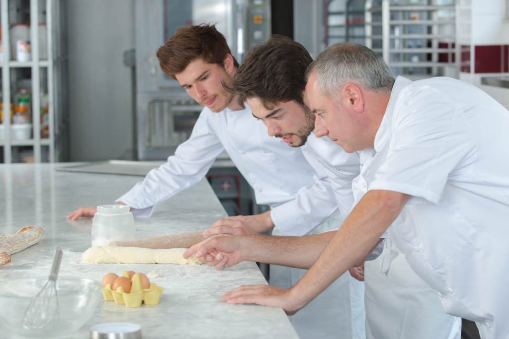 boulanger montrant une pâte sur le plan de travail à ses deux apprentis
